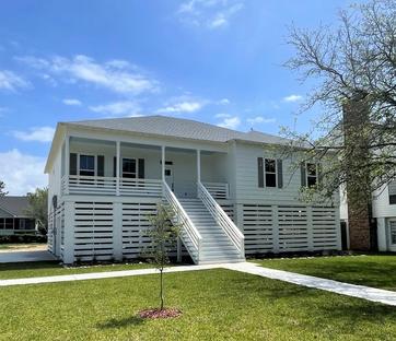 A stylish elevated home by Viridis Construction, showcasing a white coastal-inspired exterior with a metal roof, open front porch, and lattice-style skirting. The home is set on a spacious grassy lot with a sleek, minimalistic design approach.